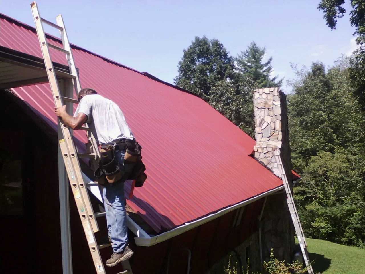 Roofer on a ladder beside a red metal roof and stone chimney — Soffit & Fascia Repair by Roofing Brothers in Menomonee Falls